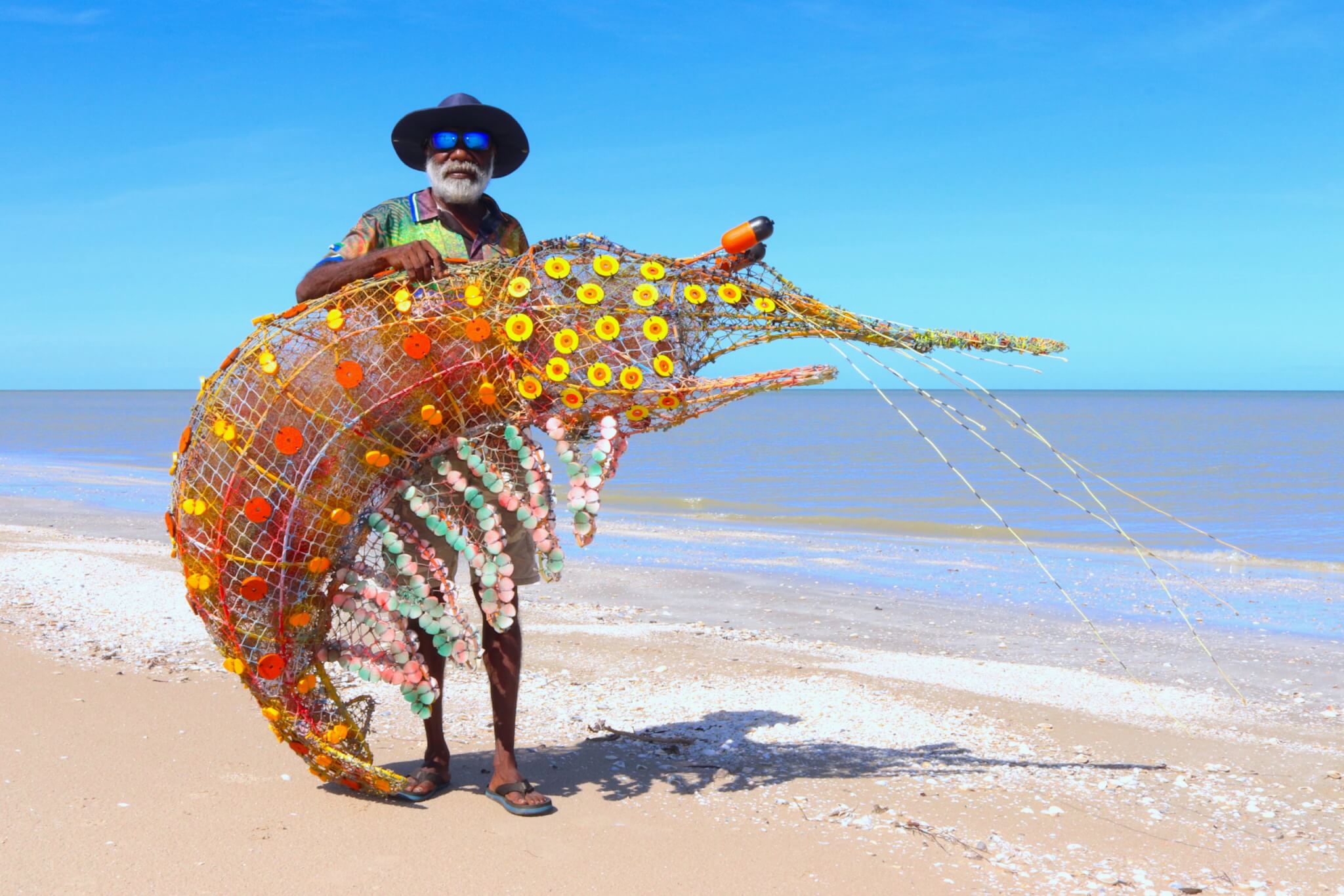 Pormpuraaw artist on a beach holding a prawn sculpture made from ghost nets
